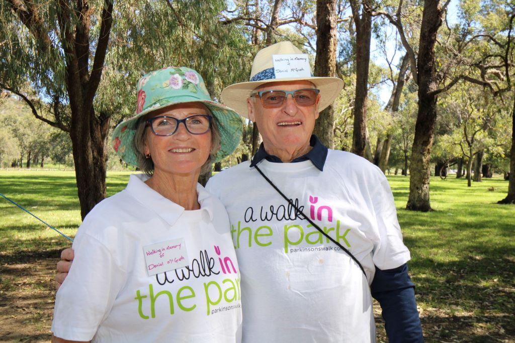 An older lady and a man smile as they stand in a park. They both wear hats and glasses.